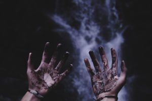 Upturned hands covered in mud or dark substance with a black and grey smoky background. Photo by Stormseeker on Unsplash